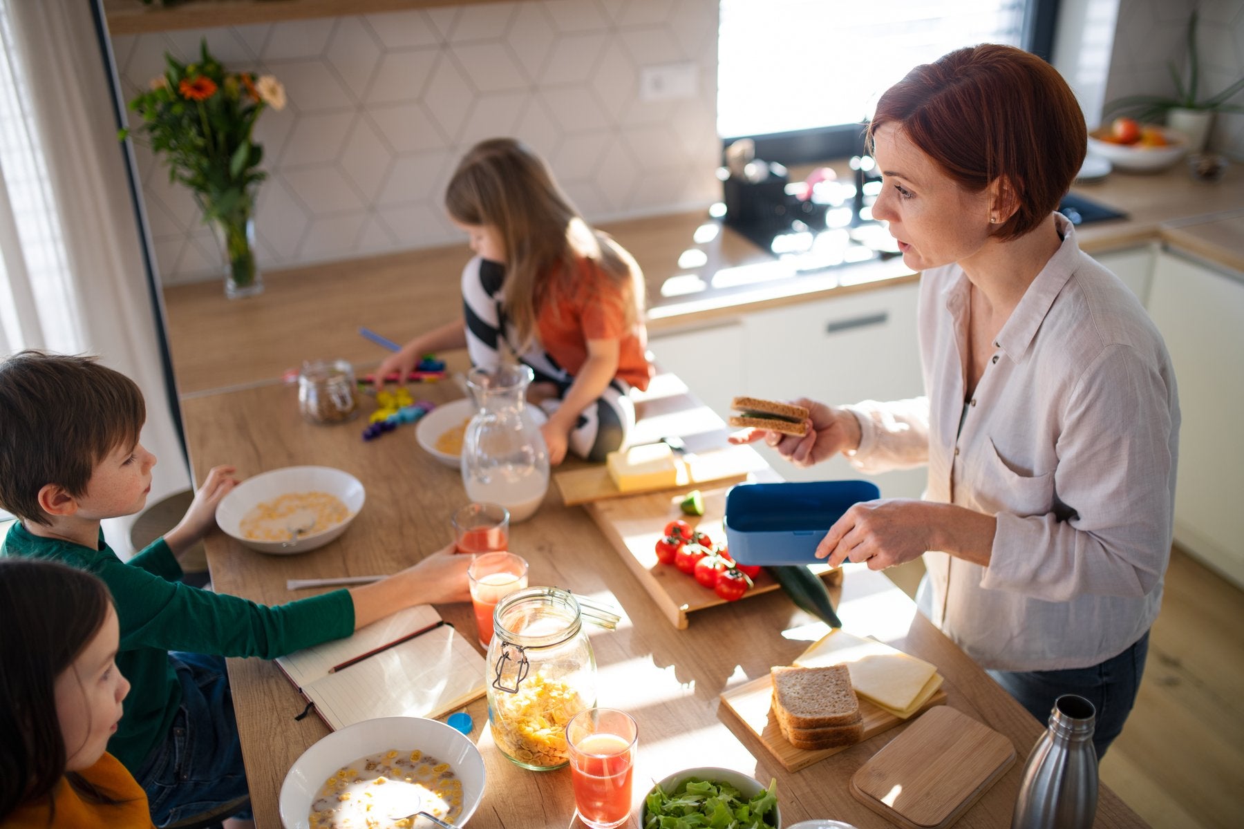 A woman preparing sandwiches in a kitchen with three children eating breakfast.