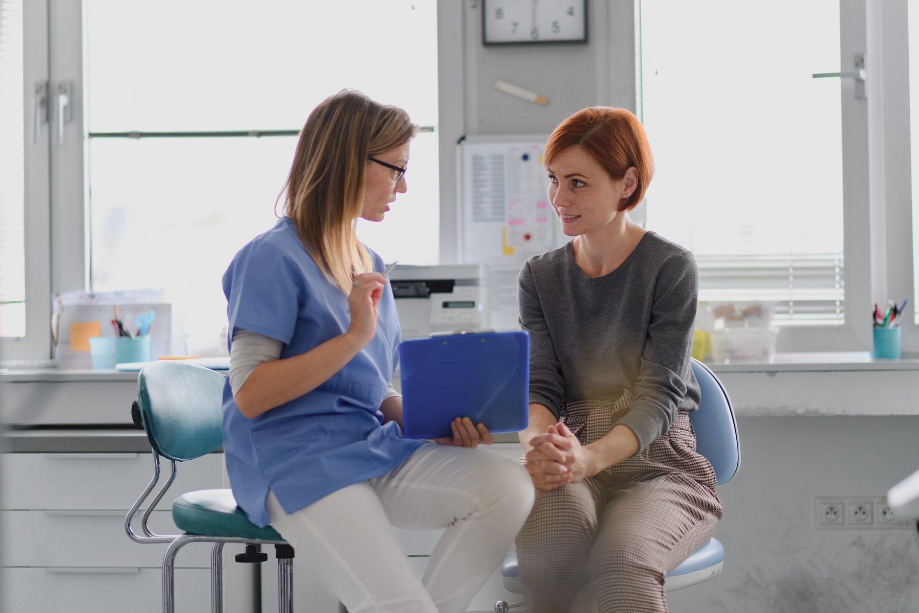 Two women sitting in a medical office, one in scrubs holding a clipboard.