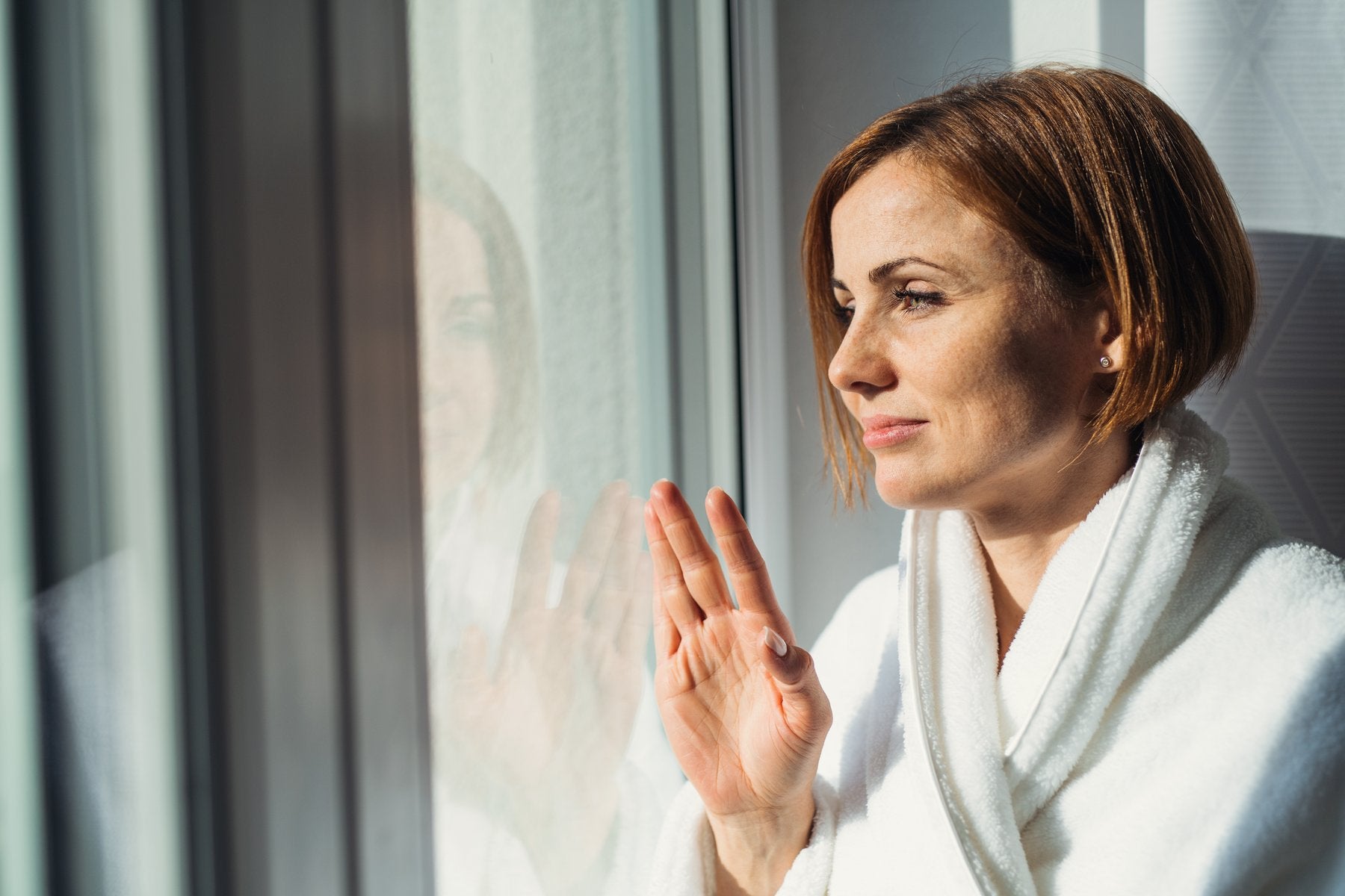 Woman in a white robe looking out a window, hand touching glass.